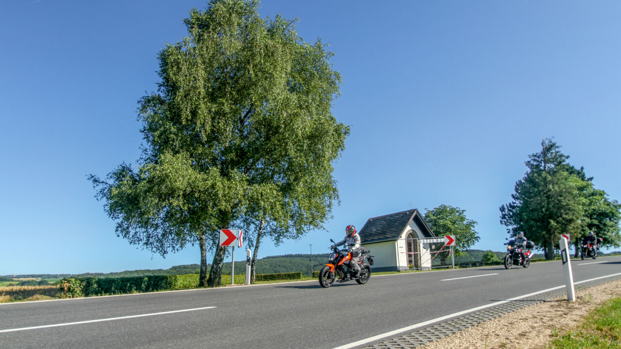 Motorräder auf einer Landstraße mit Kurvenpfeil, daneben ein kleines weißes Haus und eine Birke Motorräder auf einer Landstraße mit Kurvenpfeil, daneben ein kleines weißes Haus und eine Birke