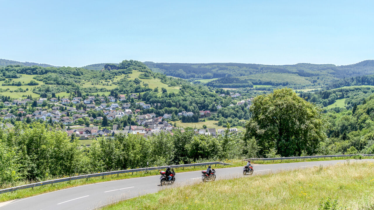 Drei Motorräder auf einer Straße am Hang, darunter ein Ort im Tal zwischen bewaldeten Kuppen Drei Motorräder auf einer Straße am Hang, darunter ein Ort im Tal zwischen bewaldeten Kuppen